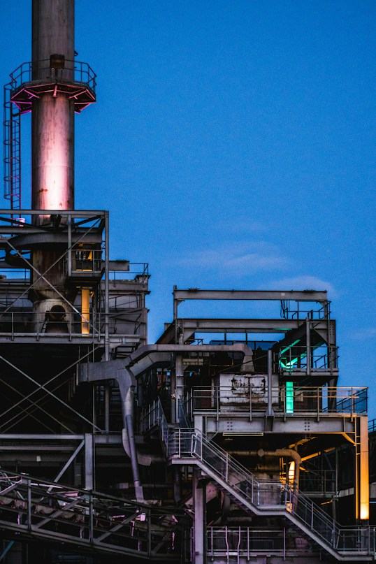 An industrial plant with stairs, platforms and a lit stack at dusk