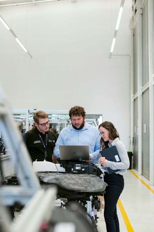 Three colleagues reviewing a laptop beside equipment on a factory floor